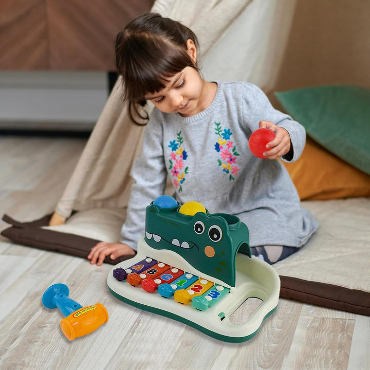 Child holding a red ball beside the alligator xylophone toy on the floor