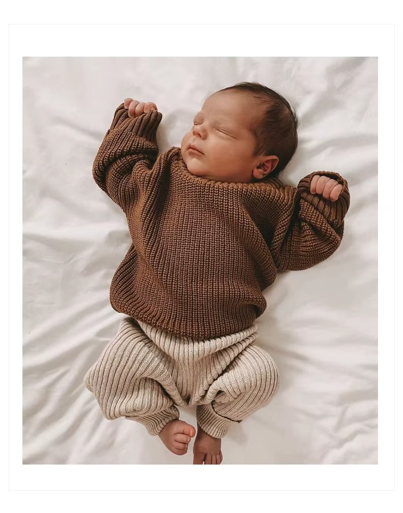 Baby lying on white bedding in a brown knit sweater and beige pants.