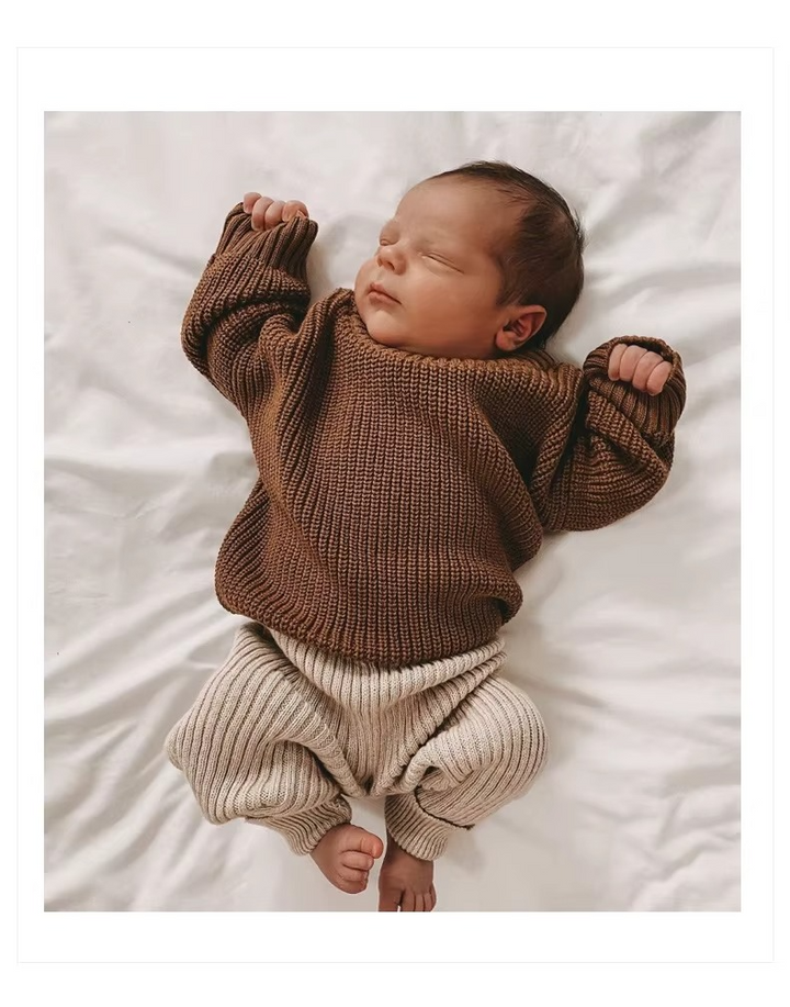 Baby lying on white bedding in a brown knit sweater and beige pants.