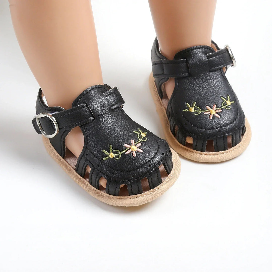 Black floral baby sandals with adjustable straps and cutout toes on mannequin feet