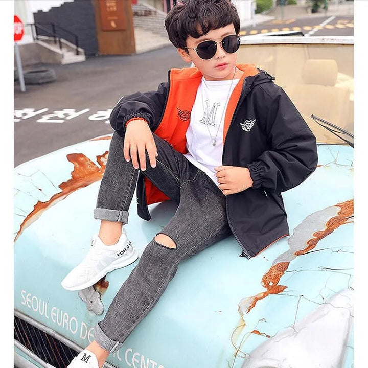 Child seated outdoors wearing a black hooded jacket with orange lining.