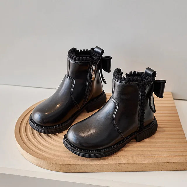 Black zip-up ankle boots with rounded toes and back bows on a wooden display