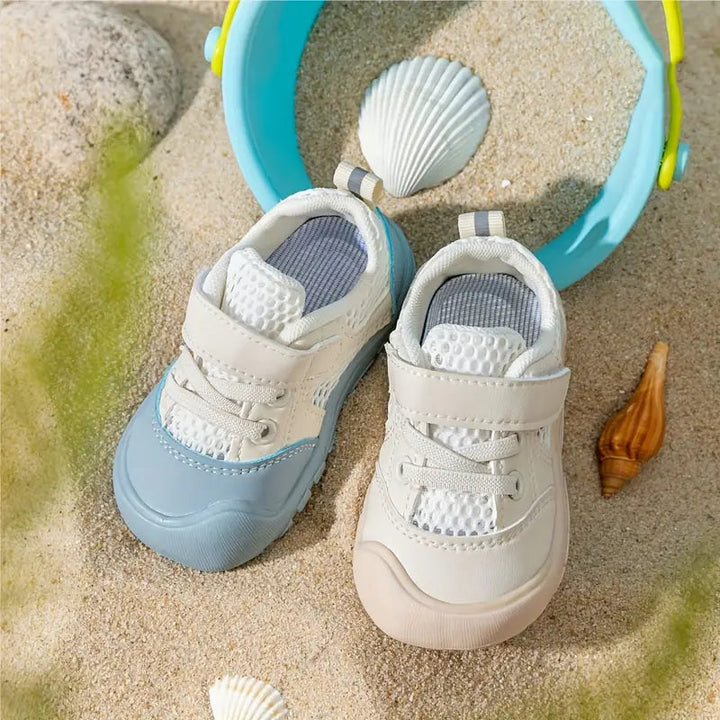 Blue and beige baby shoes viewed from above on sand beside a bucket and seashells
