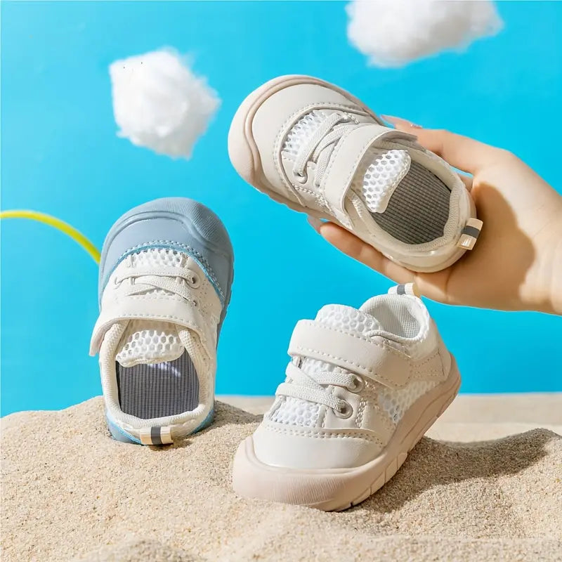 Blue and beige barefoot baby shoes displayed on sand, with a beige shoe held above them