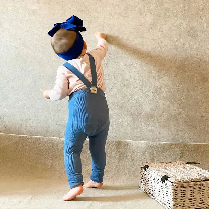 Baby wearing blue suspender leggings with a pink long-sleeve top