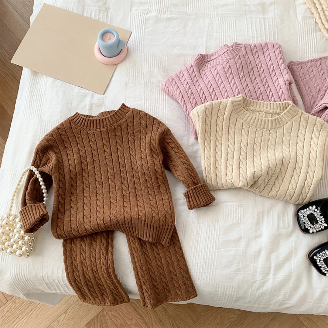 Brown cable-knit sweater and pants laid out beside folded beige and pink sweaters