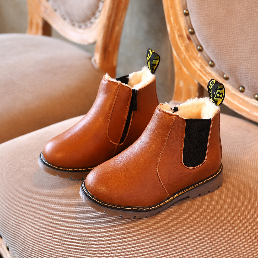Brown kids' Chelsea boots with cream-colored lining displayed on an upholstered chair