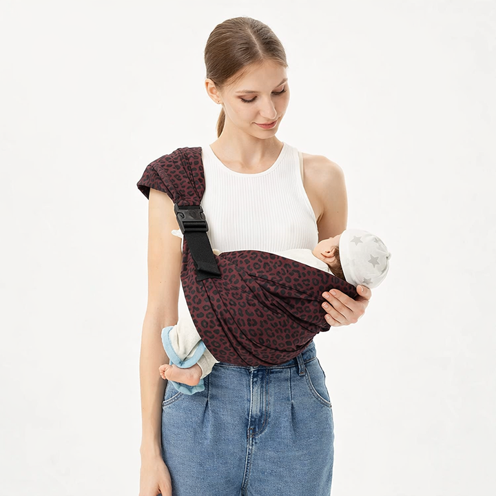 Adult supporting a child in a burgundy leopard-print sling with a wide shoulder strap