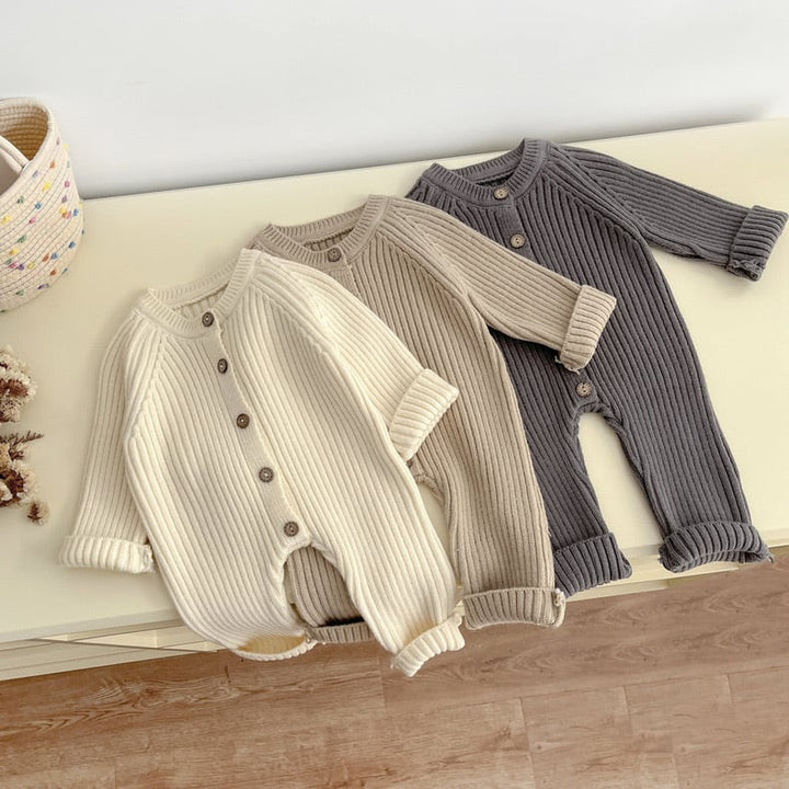 Three neutral-colored knitted baby rompers laid out with button fronts and cuffed legs