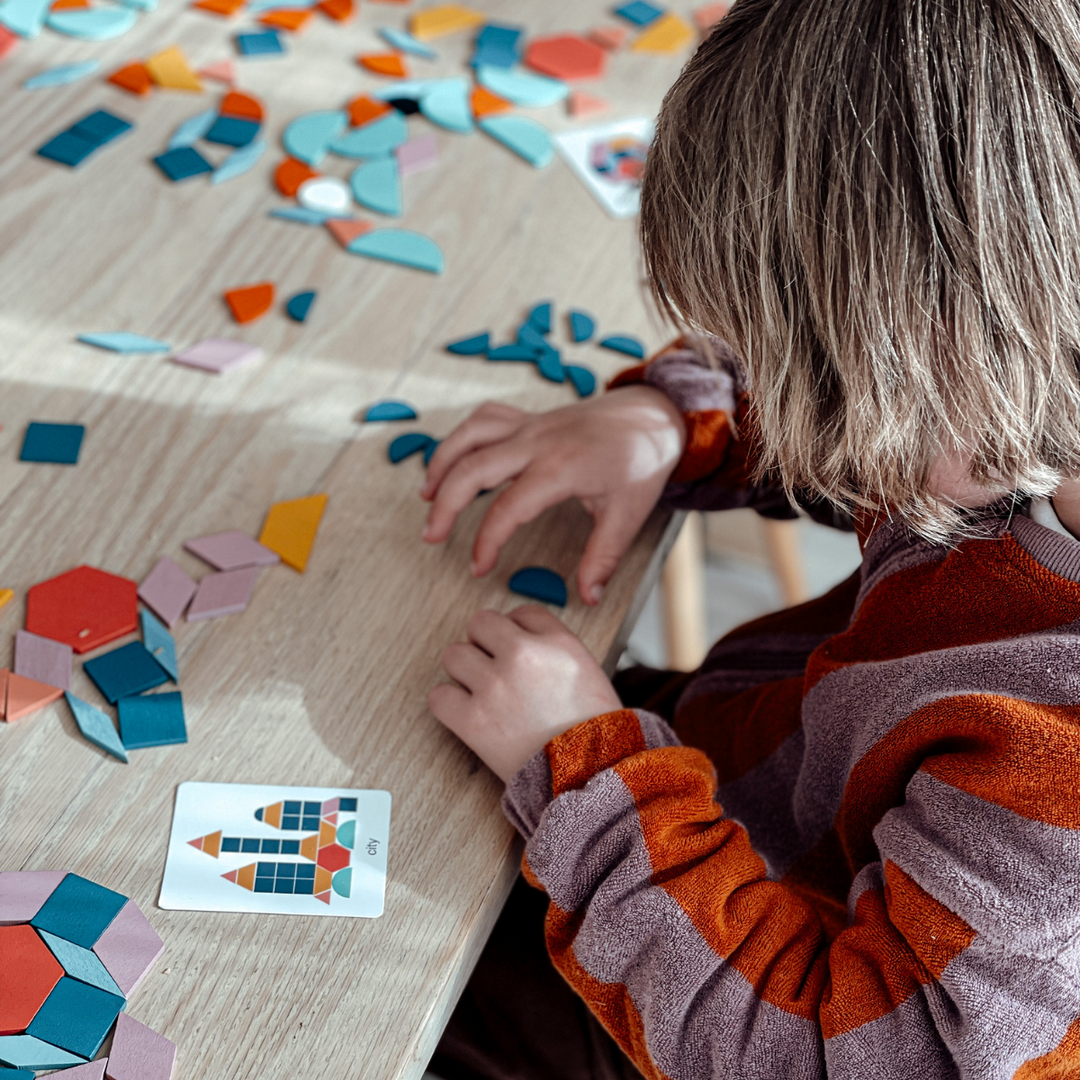 Child arranging colorful wooden shapes beside a pattern card at a table