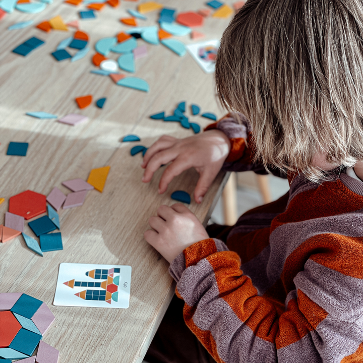 Child arranging colorful wooden shapes beside a pattern card at a table
