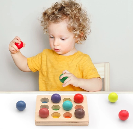 A child holds red and green balls above a wooden color-matching board