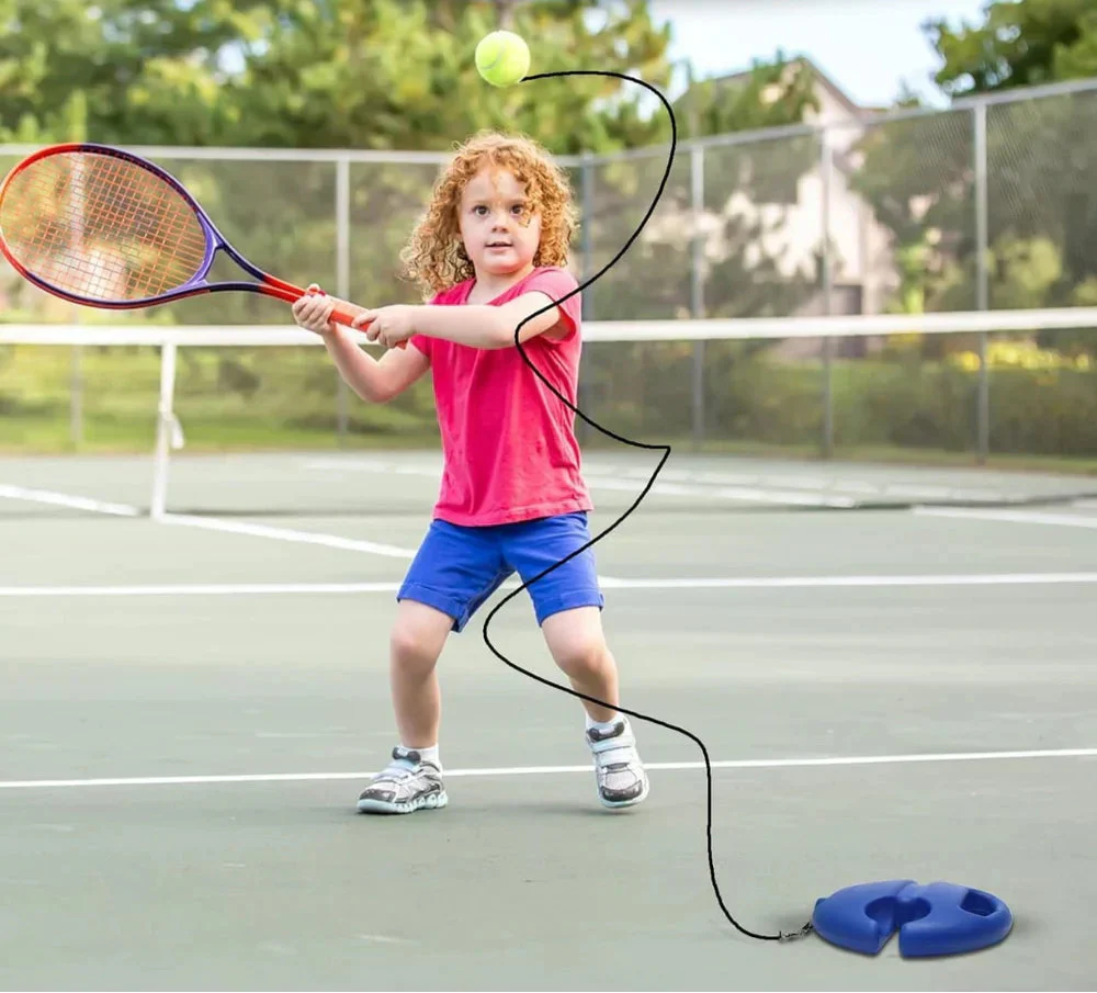 Child practicing a tennis swing on an outdoor court with a ball tethered to a blue base