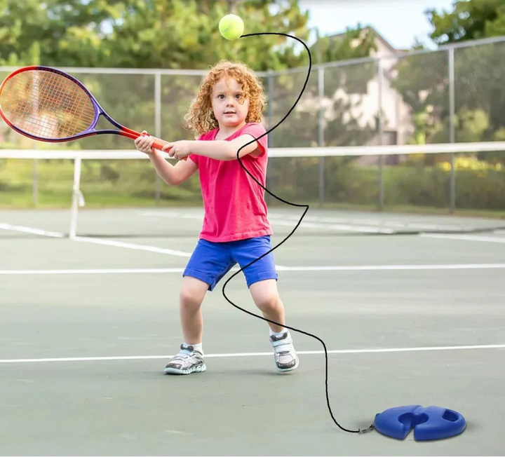Child practicing a tennis swing on an outdoor court with a ball tethered to a blue base