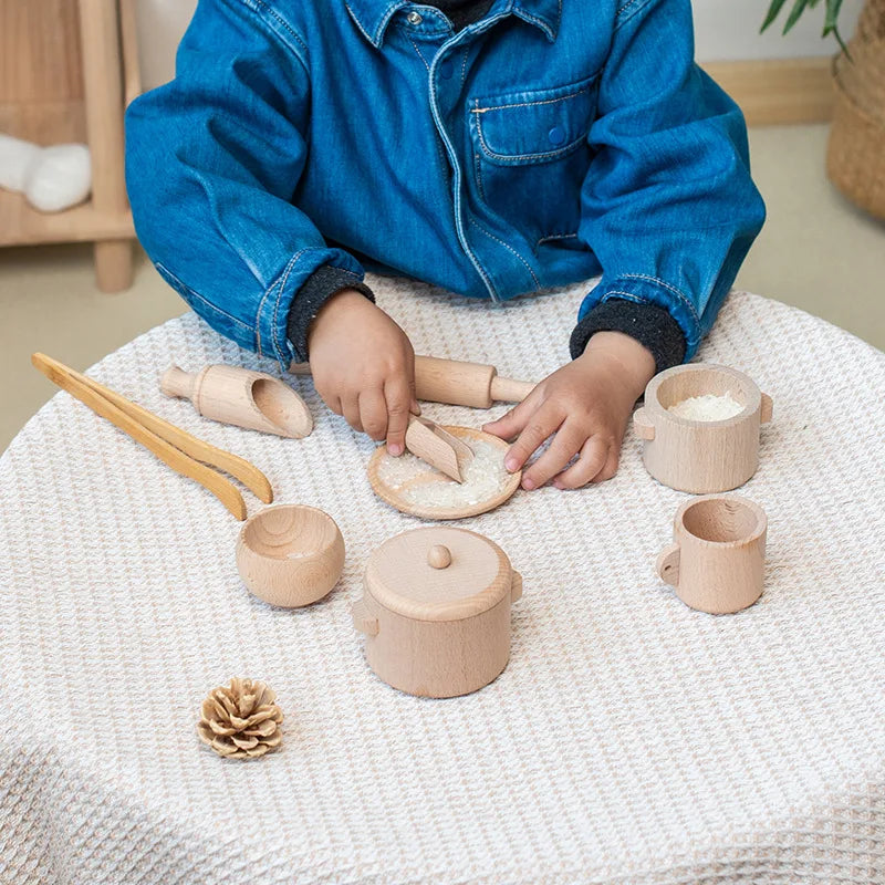 Child playing with wooden kitchen utensils, cups and a rolling pin on a cloth-covered table.
