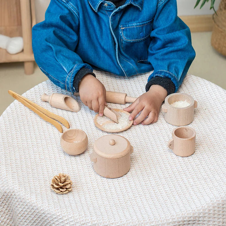 Child playing with wooden kitchen utensils, cups and a rolling pin on a cloth-covered table.