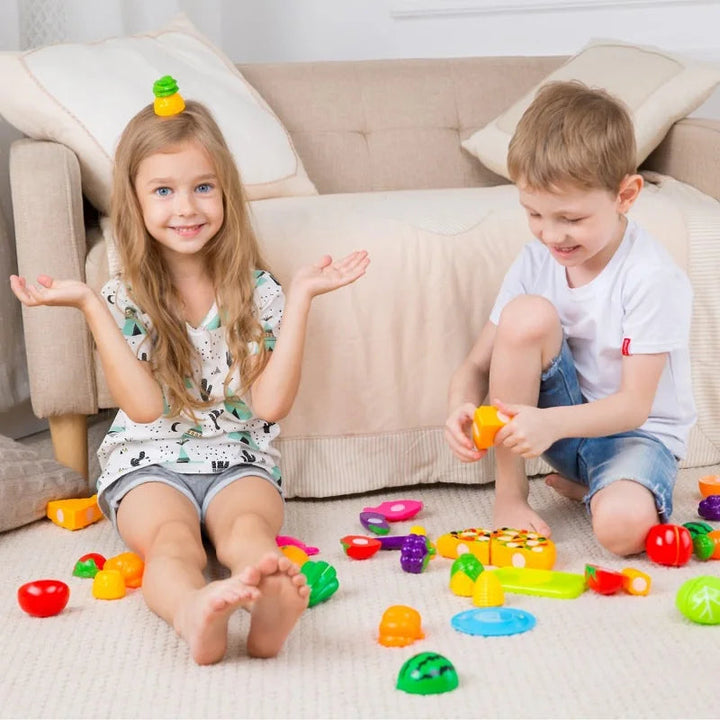 Two children playing with colorful cut-apart foods, a toy pizza, and cutting boards on the floor