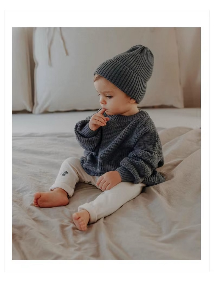 Baby sitting on a bed in a dark gray knit sweater and matching beanie.