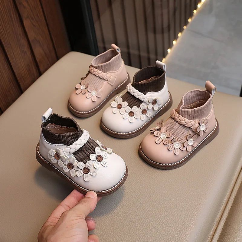 Cream White and Beige girls' ankle boots with flower appliqués and braided trim arranged on a cushioned chair.