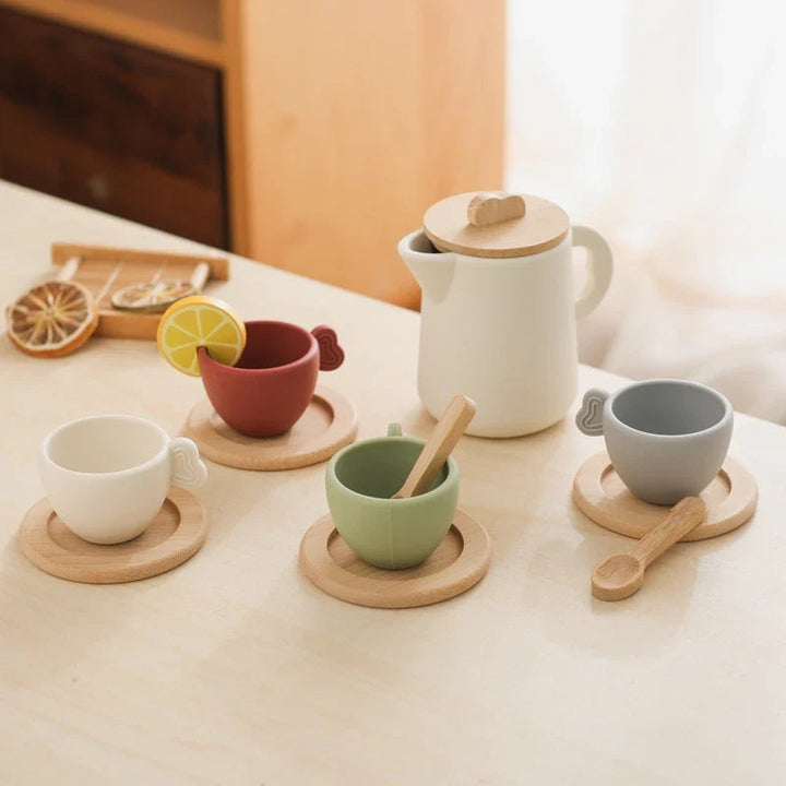 Four colored tea cups on wooden saucers beside a lidded pitcher and play spoons