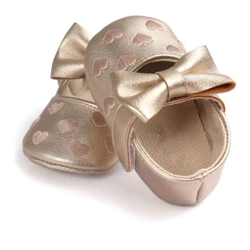 Overlapping gold baby shoes showing bow accents, heart details, and the interior