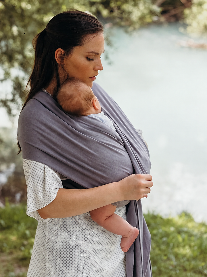 Parent wearing a gray baby wrap carrier beside the water.