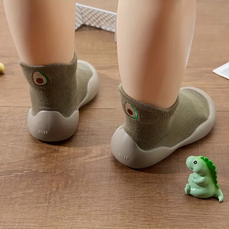 Green sock-style baby shoes worn by a child, showing avocado motifs and pale soles