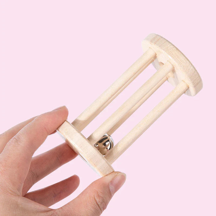 Hand holding a long wooden cage rattle with a silver-colored bell against a pink background