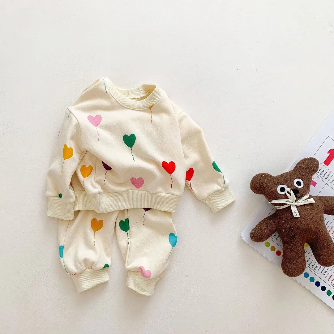 Cream baby sweater and matching heart-print pants beside a teddy bear