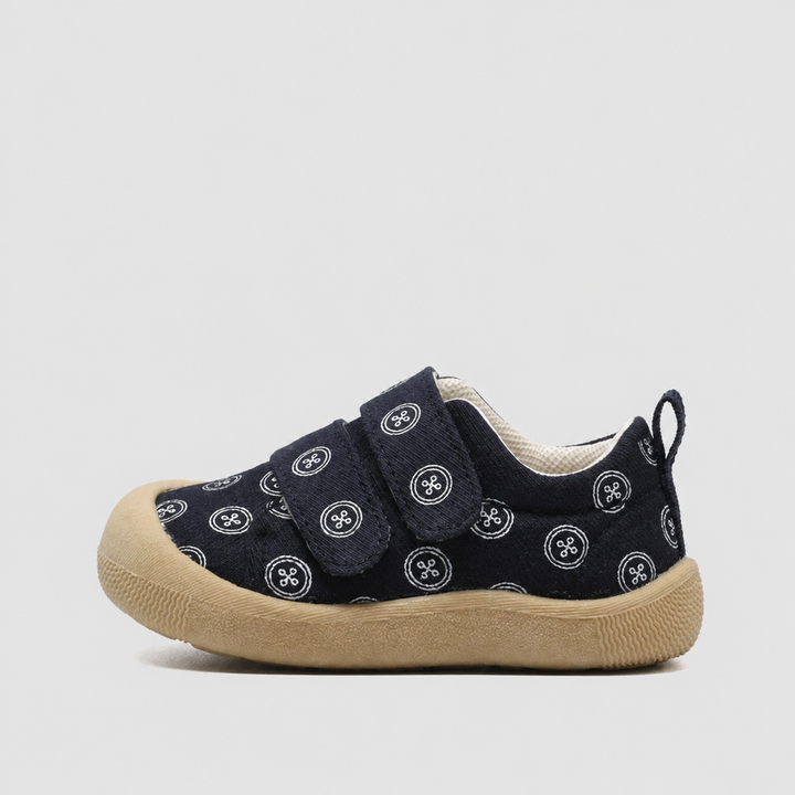 Side view of a navy canvas sneaker with circular prints, a strap closure, and a tan sole