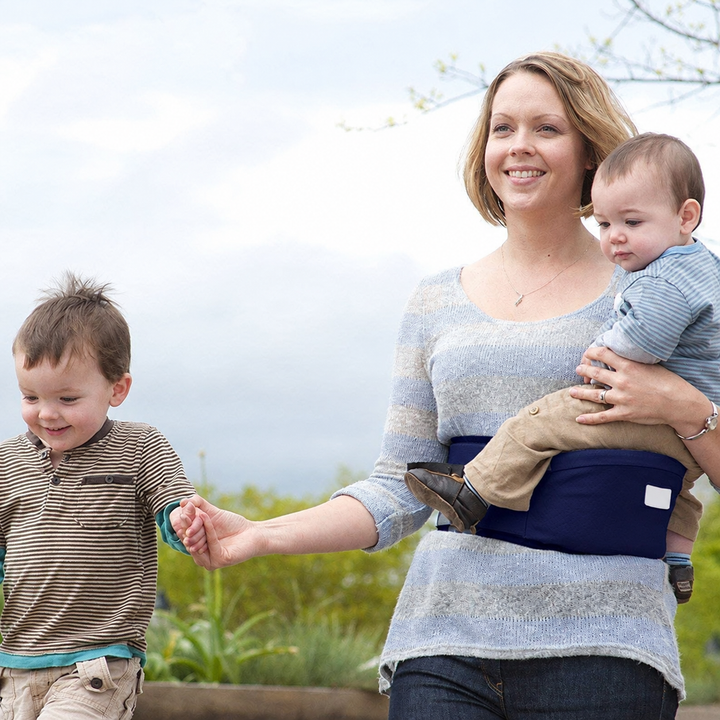 Adult holding a child on a navy hip carry seat while walking with another child outdoors