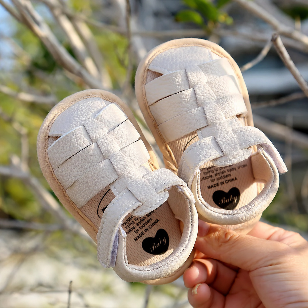 Off-white closed-toe baby sandals held outdoors, showing interwoven straps