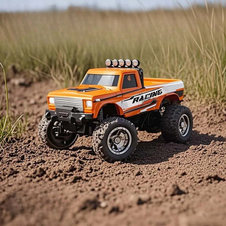 Orange mini RC pickup truck with large treaded tires on dirt beside tall grass.
