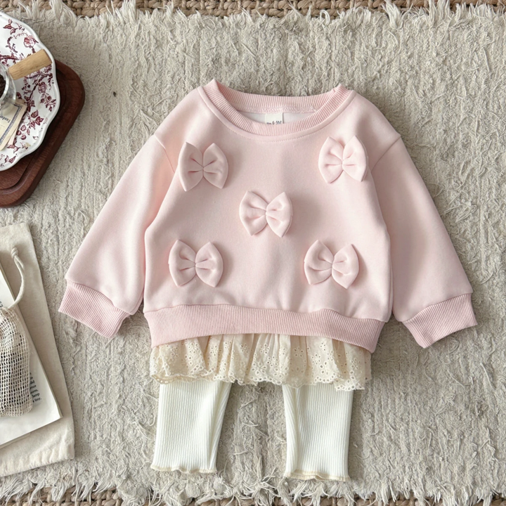 Pink baby sweater with five bows and a lace hem, paired with cream ribbed pants on a textured blanket