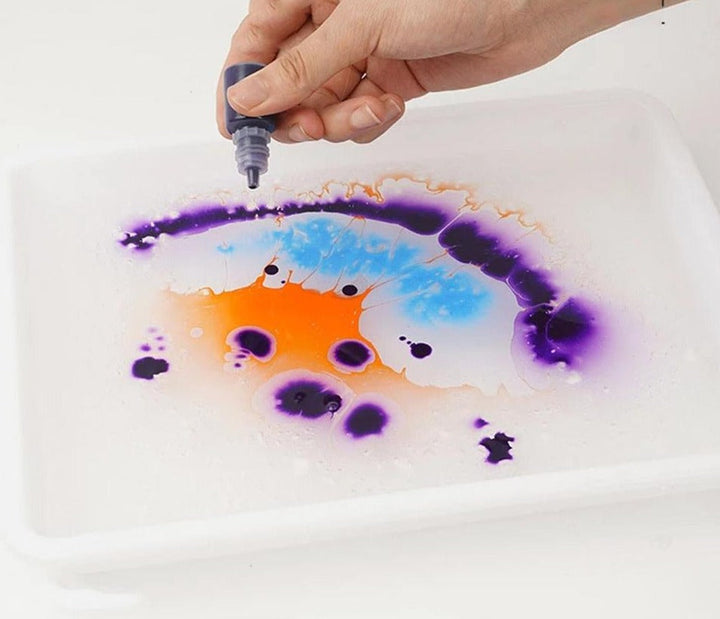 Purple, blue, and orange marbling paint being dropped onto water in a white tray