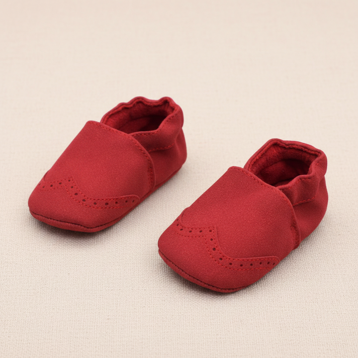 Red baby shoes with gathered ankle openings and rounded, perforated toes on beige fabric