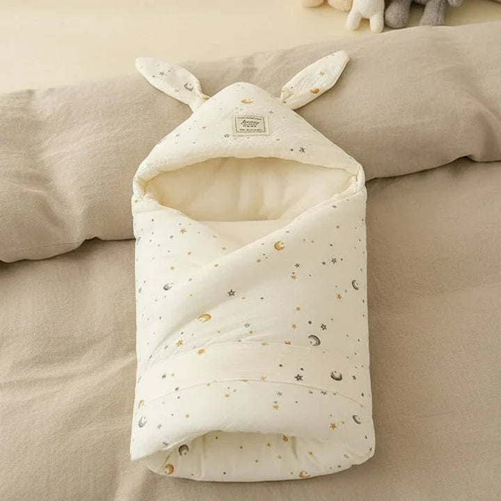 Star-and-moon print swaddle blanket with bunny ears resting on neutral bedding
