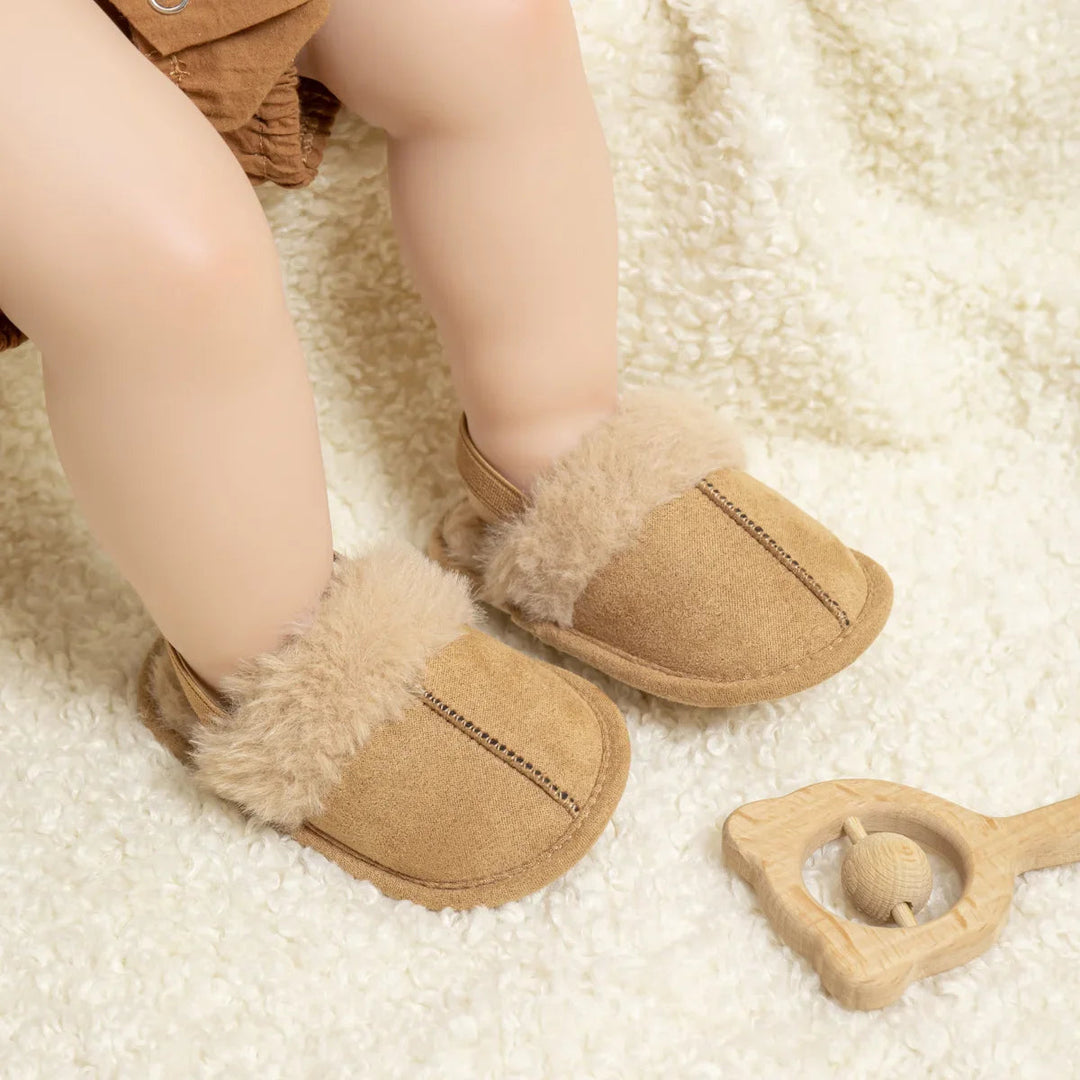 Baby wearing tan slippers with plush trim and elastic heel straps