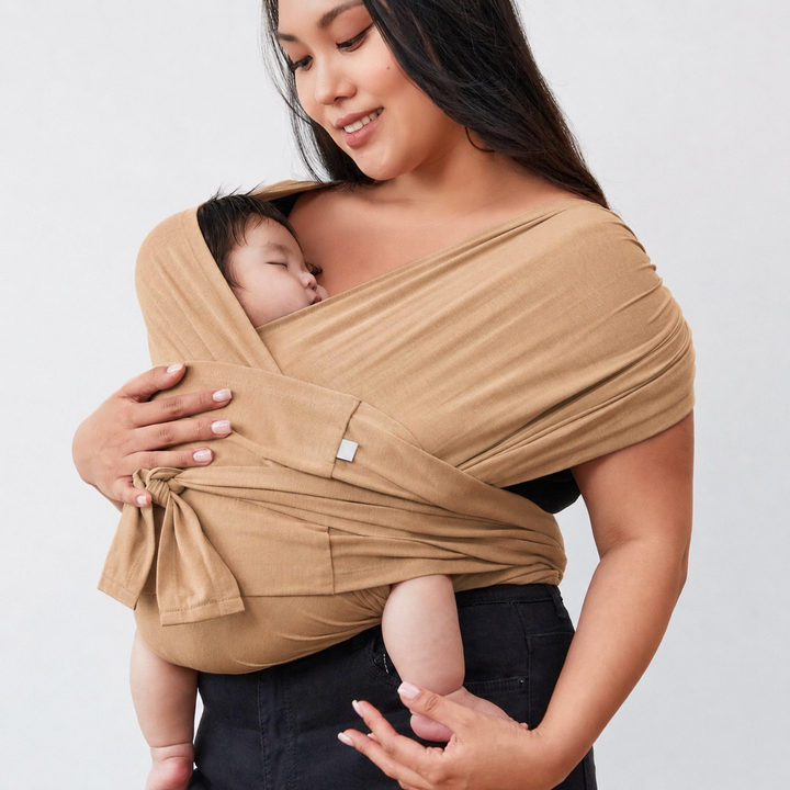 Parent holding a sleeping baby in a taupe wrap-style carrier with wide shoulder fabric