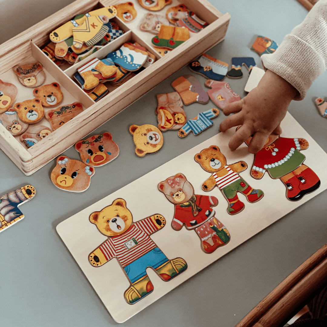 Child's hand arranging colorful bear dress-up puzzle pieces beside a wooden storage box
