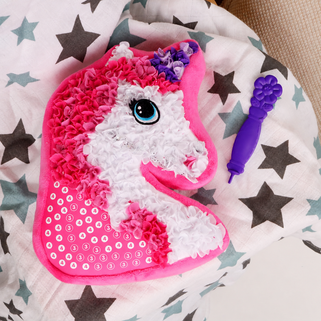 Partly decorated unicorn pillow on star-patterned bedding beside a purple crafting tool.