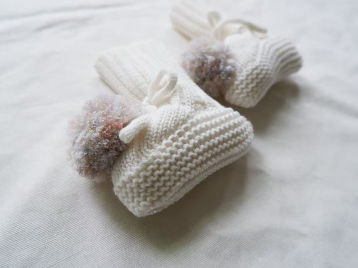 White knitted baby booties with multicolor pom-poms and ribbed cuffs.