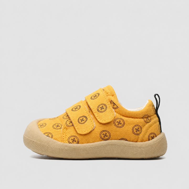 Side view of a yellow canvas sneaker with circular prints, a strap closure, and a tan sole